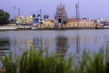 A vibrant, intricately designed temple stands prominently near a body of water, with its colorful architecture reflecting on the water's surface. Surrounding the temple are various buildings, including multi-storied structures and communication towers. Lush greenery is visible in the foreground, adding to the serenity of the scene.