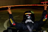 A person wearing a helmet and a racing suit raises both arms in a victory gesture. The scene is dimly lit with bright red lights forming a trail in the background, likely indicating movement or a curved path.