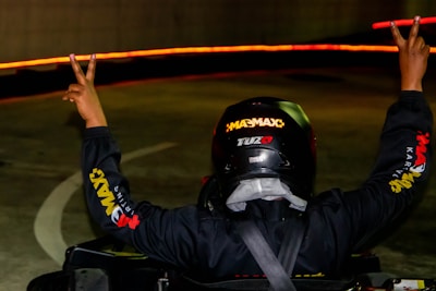 A person wearing a helmet and a racing suit raises both arms in a victory gesture. The scene is dimly lit with bright red lights forming a trail in the background, likely indicating movement or a curved path.