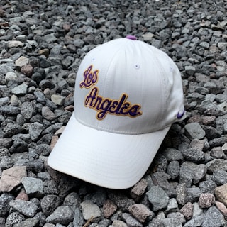 A white baseball cap with the words 'Los Angeles' embroidered in purple and golden-yellow lettering is resting on a ground covered with small gray and brown stones. The cap features a purple Nike logo on the side.