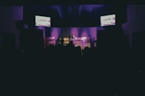 A dimly lit stage features a solo performer standing under spotlights, with purple lighting creating an atmospheric background. Two screens on either side display abstract images, adding a modern touch. A darkened audience can be seen in the foreground.