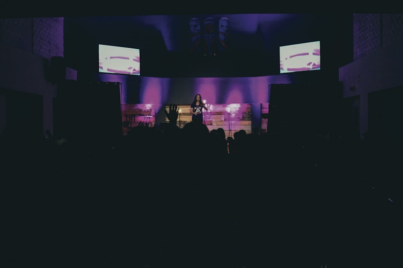 A dimly lit stage features a solo performer standing under spotlights, with purple lighting creating an atmospheric background. Two screens on either side display abstract images, adding a modern touch. A darkened audience can be seen in the foreground.