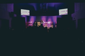 A dimly lit stage features a solo performer standing under spotlights, with purple lighting creating an atmospheric background. Two screens on either side display abstract images, adding a modern touch. A darkened audience can be seen in the foreground.