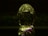 A decorative, transparent crystal object shaped like an egg with intricate carvings and patterns reflecting light. It features subtle iridescent colors on a dark background, and the object is mirrored against a surface.