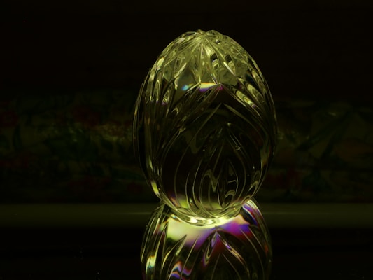A decorative, transparent crystal object shaped like an egg with intricate carvings and patterns reflecting light. It features subtle iridescent colors on a dark background, and the object is mirrored against a surface.