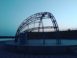 A metal framework structure is situated outdoors on a circular paved platform. The surrounding area includes tiered seating, likely for an audience, and the scene is illuminated by ambient twilight. The sky is clear with a gradient of light blue tones, and a body of water is visible in the background.