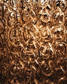 A dense cluster of translucent, intertwined acrylic chains with a golden hue, creating an intricate and luxurious texture. The light shines through them, adding a warm, glowing effect.