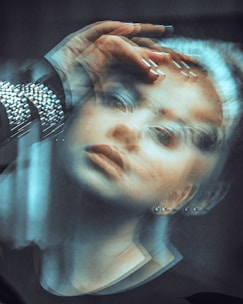 A double exposure effect creates a surreal and ethereal image of a woman with her hand over her forehead. The composition is blurred and layered, giving a sense of movement or dreamlike quality. Her face and jewelry, including earrings and a metallic bracelet, are partially visible.