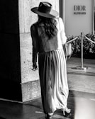A person wearing a long pleated skirt, a textured jacket, and a wide-brimmed hat is walking towards the entrance of a store labeled 'Dior Millefiori Garden'. The picture is in black and white and the setting seems to be an upscale outdoor shopping area, with a polished stone surface and a decorative rope barrier in the scene.