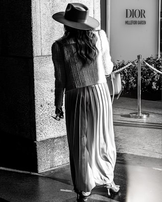 A person wearing a long pleated skirt, a textured jacket, and a wide-brimmed hat is walking towards the entrance of a store labeled 'Dior Millefiori Garden'. The picture is in black and white and the setting seems to be an upscale outdoor shopping area, with a polished stone surface and a decorative rope barrier in the scene.