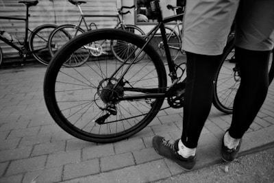 A person in cycling attire stands next to a bicycle on a paved surface. Several other bicycles are parked nearby, leaning against a wall. The focus is on the rear wheel and gear mechanism of the bicycle.