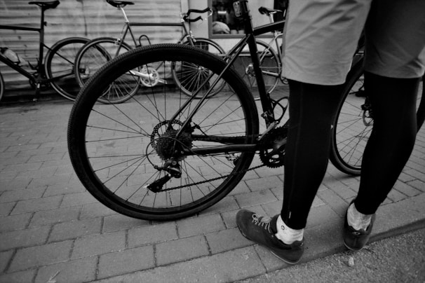 A person in cycling attire stands next to a bicycle on a paved surface. Several other bicycles are parked nearby, leaning against a wall. The focus is on the rear wheel and gear mechanism of the bicycle.