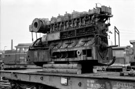 A large industrial engine sits on a flatbed freight car in a train yard, surrounded by other trains and industrial equipment. The engine appears aged with visible rust and wear, and is mounted on wooden logs. Power lines and a smokestack can be seen in the background.
