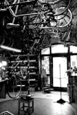 A vintage bicycle workshop filled with various bicycle frames and parts hanging from the ceiling. The room has a rustic and cluttered appearance with tools and equipment scattered around. A bicycle is mounted on a stand for repairs in the center of the room, and the walls are lined with shelves holding more parts and tools. Light comes in through a window at the back, partially illuminating the space.