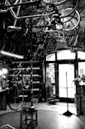 A vintage bicycle workshop filled with various bicycle frames and parts hanging from the ceiling. The room has a rustic and cluttered appearance with tools and equipment scattered around. A bicycle is mounted on a stand for repairs in the center of the room, and the walls are lined with shelves holding more parts and tools. Light comes in through a window at the back, partially illuminating the space.
