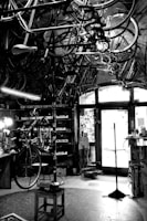 A vintage bicycle workshop filled with various bicycle frames and parts hanging from the ceiling. The room has a rustic and cluttered appearance with tools and equipment scattered around. A bicycle is mounted on a stand for repairs in the center of the room, and the walls are lined with shelves holding more parts and tools. Light comes in through a window at the back, partially illuminating the space.