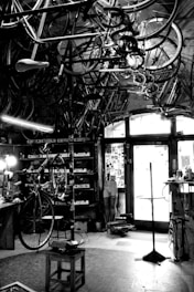 A vintage bicycle workshop filled with various bicycle frames and parts hanging from the ceiling. The room has a rustic and cluttered appearance with tools and equipment scattered around. A bicycle is mounted on a stand for repairs in the center of the room, and the walls are lined with shelves holding more parts and tools. Light comes in through a window at the back, partially illuminating the space.