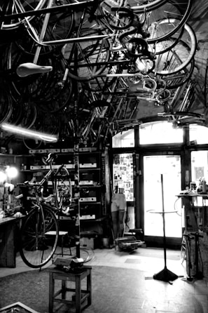 A vintage bicycle workshop filled with various bicycle frames and parts hanging from the ceiling. The room has a rustic and cluttered appearance with tools and equipment scattered around. A bicycle is mounted on a stand for repairs in the center of the room, and the walls are lined with shelves holding more parts and tools. Light comes in through a window at the back, partially illuminating the space.