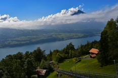 A picturesque landscape features a serene lake surrounded by lush greenery and rolling hills. A mountain peak is partially shrouded by clouds, rising in the background under a blue sky. A few wooden houses with red roofs are nestled among the trees, adding a rustic charm to the scene.