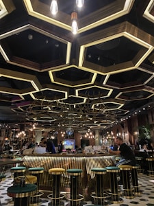An upscale bar with a modern, geometric ceiling design featuring hexagonal light fixtures and metallic accents. The bar counter is surrounded by tall stools with green and yellow cushions. Warm lighting creates a cozy atmosphere, and there are multiple patrons engaged in conversation and activity.