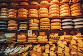 An assortment of large, round cheese wheels are neatly stacked on wooden shelves, creating a wall of cheese. The wheels come in different shades of yellow and are labeled with various brand names. Smaller cheese pieces are displayed on the lower shelves.