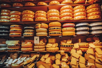 An assortment of large, round cheese wheels are neatly stacked on wooden shelves, creating a wall of cheese. The wheels come in different shades of yellow and are labeled with various brand names. Smaller cheese pieces are displayed on the lower shelves.
