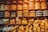 An assortment of large, round cheese wheels are neatly stacked on wooden shelves, creating a wall of cheese. The wheels come in different shades of yellow and are labeled with various brand names. Smaller cheese pieces are displayed on the lower shelves.