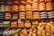 An assortment of large, round cheese wheels are neatly stacked on wooden shelves, creating a wall of cheese. The wheels come in different shades of yellow and are labeled with various brand names. Smaller cheese pieces are displayed on the lower shelves.