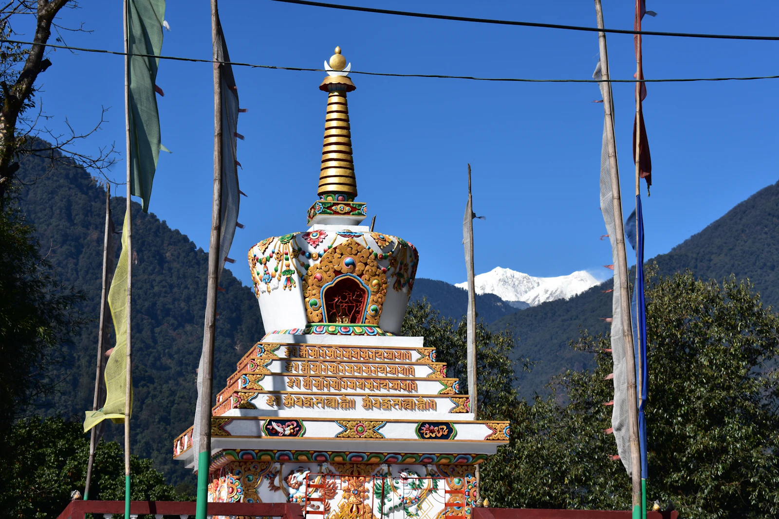 A painted stupa and tall prayer flags in Yuksom, with the snow summit of Kabru rising at the head of the valley