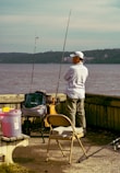 A person stands by the water with fishing gear, including two fishing rods and various supplies on a cart. They are surrounded by a bench and a folding chair, while wearing casual outdoor clothing and a cap. The background features a body of water with hills on the opposite shore.