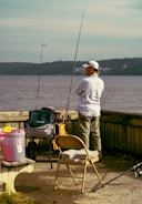 A person stands by the water with fishing gear, including two fishing rods and various supplies on a cart. They are surrounded by a bench and a folding chair, while wearing casual outdoor clothing and a cap. The background features a body of water with hills on the opposite shore.