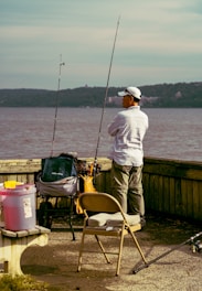 A person stands by the water with fishing gear, including two fishing rods and various supplies on a cart. They are surrounded by a bench and a folding chair, while wearing casual outdoor clothing and a cap. The background features a body of water with hills on the opposite shore.