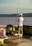 A person stands by the water with fishing gear, including two fishing rods and various supplies on a cart. They are surrounded by a bench and a folding chair, while wearing casual outdoor clothing and a cap. The background features a body of water with hills on the opposite shore.