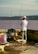 A person stands by the water with fishing gear, including two fishing rods and various supplies on a cart. They are surrounded by a bench and a folding chair, while wearing casual outdoor clothing and a cap. The background features a body of water with hills on the opposite shore.