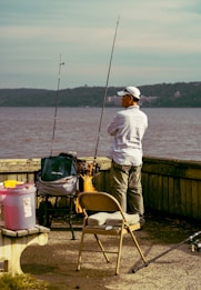 A person stands by the water with fishing gear, including two fishing rods and various supplies on a cart. They are surrounded by a bench and a folding chair, while wearing casual outdoor clothing and a cap. The background features a body of water with hills on the opposite shore.