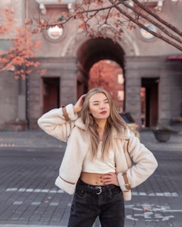 A young woman with long hair stands confidently on a cobblestone street. She is wearing a white fluffy jacket, a crop top, and black jeans. The background features an architectural archway and trees with autumn leaves, creating a warm and inviting urban scene.