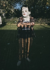A person is standing outdoors in a dimly lit environment, wearing a Halloween mask resembling a classic horror character with green skin and scars. They are dressed casually in a plaid shirt over a t-shirt with lettering, and checkered slip-on shoes. The backdrop consists of a grass lawn, a chain-link fence, and abundant vegetation in the shadows.