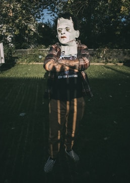 A person is standing outdoors in a dimly lit environment, wearing a Halloween mask resembling a classic horror character with green skin and scars. They are dressed casually in a plaid shirt over a t-shirt with lettering, and checkered slip-on shoes. The backdrop consists of a grass lawn, a chain-link fence, and abundant vegetation in the shadows.