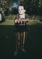 A person is standing outdoors in a dimly lit environment, wearing a Halloween mask resembling a classic horror character with green skin and scars. They are dressed casually in a plaid shirt over a t-shirt with lettering, and checkered slip-on shoes. The backdrop consists of a grass lawn, a chain-link fence, and abundant vegetation in the shadows.