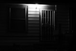 A dimly lit exterior door and window on a house at night, with a security grille over the door, illuminated by a single overhead light, casting shadows on the siding.