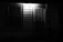 A dimly lit exterior door and window on a house at night, with a security grille over the door, illuminated by a single overhead light, casting shadows on the siding.