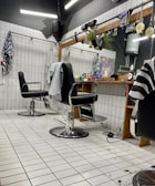 The image depicts an interior of a barbershop with two black leather barber chairs equipped with metal armrests and a footrest. The floor is covered with white tiles, and some hair clippings are visible. Behind the chairs is a large mirror mounted on a wooden frame, with a shelf holding various hair products and a bottle. There are hairdressing capes hanging on hooks on the tiled walls, and soft lighting gives the space a cozy appearance.