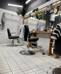 The image depicts an interior of a barbershop with two black leather barber chairs equipped with metal armrests and a footrest. The floor is covered with white tiles, and some hair clippings are visible. Behind the chairs is a large mirror mounted on a wooden frame, with a shelf holding various hair products and a bottle. There are hairdressing capes hanging on hooks on the tiled walls, and soft lighting gives the space a cozy appearance.