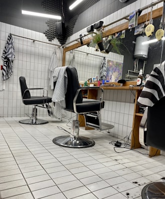 The image depicts an interior of a barbershop with two black leather barber chairs equipped with metal armrests and a footrest. The floor is covered with white tiles, and some hair clippings are visible. Behind the chairs is a large mirror mounted on a wooden frame, with a shelf holding various hair products and a bottle. There are hairdressing capes hanging on hooks on the tiled walls, and soft lighting gives the space a cozy appearance.