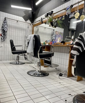 The image depicts an interior of a barbershop with two black leather barber chairs equipped with metal armrests and a footrest. The floor is covered with white tiles, and some hair clippings are visible. Behind the chairs is a large mirror mounted on a wooden frame, with a shelf holding various hair products and a bottle. There are hairdressing capes hanging on hooks on the tiled walls, and soft lighting gives the space a cozy appearance.