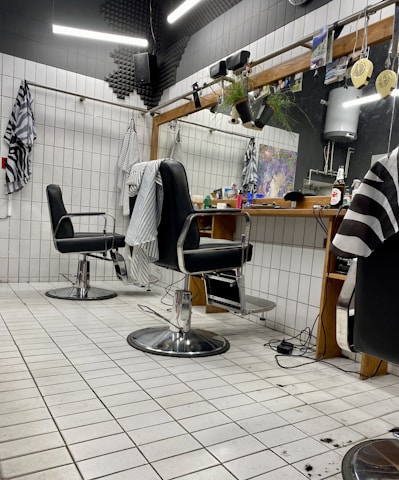 The image depicts an interior of a barbershop with two black leather barber chairs equipped with metal armrests and a footrest. The floor is covered with white tiles, and some hair clippings are visible. Behind the chairs is a large mirror mounted on a wooden frame, with a shelf holding various hair products and a bottle. There are hairdressing capes hanging on hooks on the tiled walls, and soft lighting gives the space a cozy appearance.