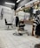 The image depicts an interior of a barbershop with two black leather barber chairs equipped with metal armrests and a footrest. The floor is covered with white tiles, and some hair clippings are visible. Behind the chairs is a large mirror mounted on a wooden frame, with a shelf holding various hair products and a bottle. There are hairdressing capes hanging on hooks on the tiled walls, and soft lighting gives the space a cozy appearance.