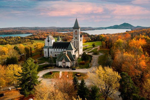 An impressive stone church with a tall spire is surrounded by vibrant autumn foliage, with a vast landscape of trees and a lake in the background. The setting sun casts a warm glow over the scene, highlighting the colorful leaves and the church's architecture.
