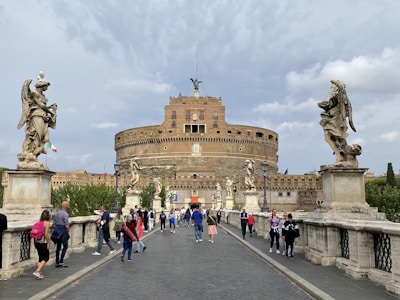 A large stone building, resembling a fortress or castle, with multiple levels and small windows. Statues of angels are positioned on either side of a bridge crowded with people. The sky is overcast, and a small Italian flag can be seen among the trees. The scene conveys a sense of historic architecture and tourism.
