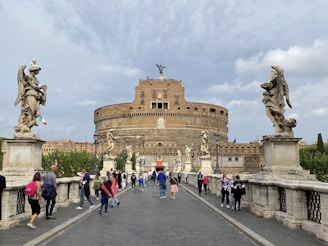 A large stone building, resembling a fortress or castle, with multiple levels and small windows. Statues of angels are positioned on either side of a bridge crowded with people. The sky is overcast, and a small Italian flag can be seen among the trees. The scene conveys a sense of historic architecture and tourism.